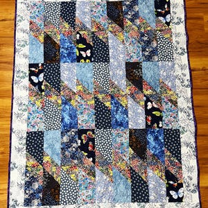 Peut inclure: Une courtepointe avec un motif de patchwork composé de différents tissus bleus et floraux. La courtepointe est bordée d'une bordure blanche et d'une garniture violette.