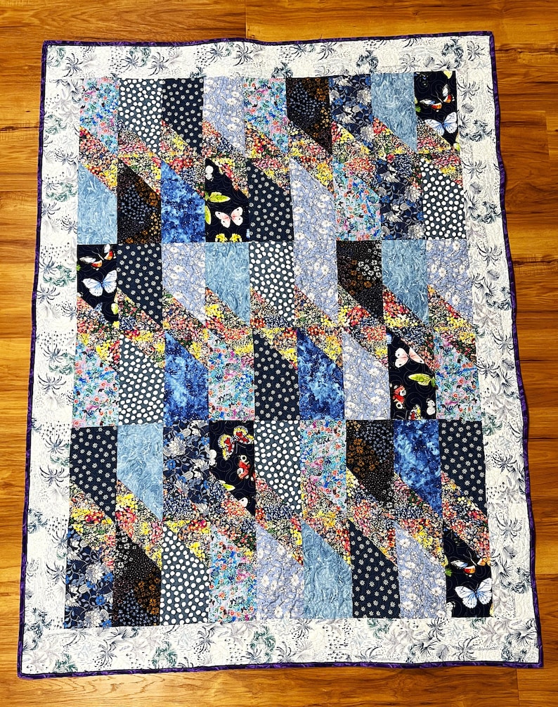 K&ouml;nnte beinhalten: Eine Steppdecke mit Patchwork-Muster aus verschiedenen blauen und floralen Stoffen. Die Decke ist mit einem wei&szlig;en Rand und einem violetten Besatz eingefasst.