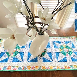 May include: A white ceramic vase with a unique design holds a bouquet of white magnolia blossoms. The vase is sitting on a table runner with a patchwork design featuring blue, green, and white fabric squares. The table runner has a green border with a floral pattern.