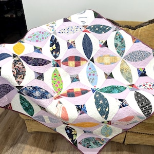 May include: A handmade quilt with a pale purple background and a repeating pattern of colourful, oval-shaped patches. The patches feature various floral, paisley, and geometric designs in shades of blue, green, red, and yellow. The quilt is draped over a brown sofa.