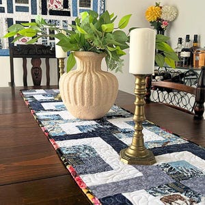 May include: A dark wood dining table is set with a blue and white patterned table runner. A beige vase with greenery and a brass candle holder with a white candle are centered on the table. A quilt hangs on the wall.