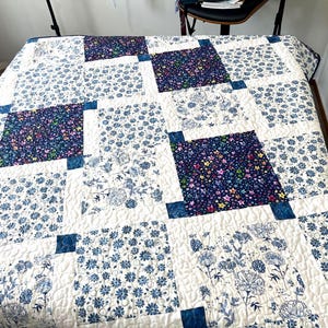 Puede incluir: Una colcha blanca con un diseño de patchwork que presenta patrones florales azules y blancos. La colcha está compuesta por nueve cuadrados, cada uno con un patrón diferente. La colcha está colocada sobre una cama.