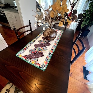 May include: A quilted table runner with a geometric pattern in shades of brown, blue, green, yellow, and white. The runner is placed on a dark wood table with a vase of dried flowers in the center.