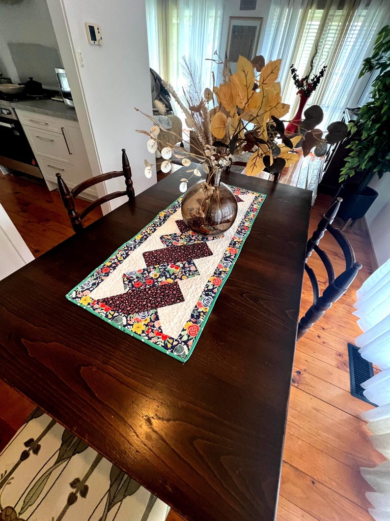 Op de afbeelding: Een gestikte tafelrunner met een geometrisch patroon in tinten bruin, blauw, groen, geel en wit. De runner ligt op een donkerhouten tafel met een vaas met gedroogde bloemen in het midden.