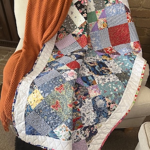 May include: A patchwork quilt featuring a mix of floral and geometric patterns in shades of blue, red, green, and white. The quilt is folded and draped over a chair with a brown throw blanket.