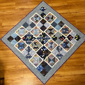 K&ouml;nnte beinhalten: Eine blaue und wei&szlig;e Steppdecke mit einem geometrischen Muster aus Quadraten und Dreiecken. Die Steppdecke ist mit einem violetten Rand eingefasst und hat einen wei&szlig;en Hintergrund mit kleinen wei&szlig;en Punkten. Die Steppdecke zeigt verschiedene Blumen- und Schmetterlingsmuster.