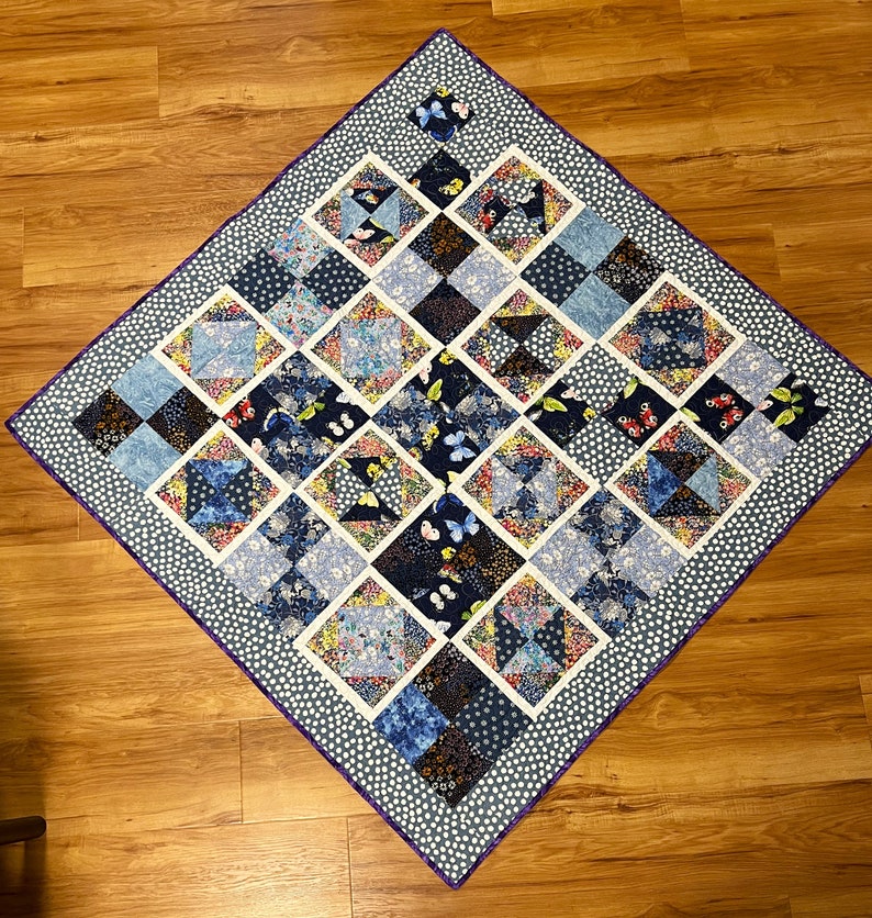 K&ouml;nnte beinhalten: Eine blaue und wei&szlig;e Steppdecke mit einem geometrischen Muster aus Quadraten und Dreiecken. Die Steppdecke ist mit einem violetten Rand eingefasst und hat einen wei&szlig;en Hintergrund mit kleinen wei&szlig;en Punkten. Die Steppdecke zeigt verschiedene Blumen- und Schmetterlingsmuster.