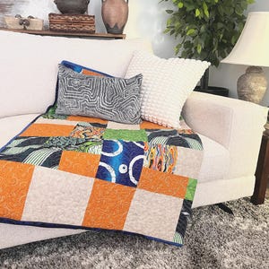 May include: A rectangular patchwork quilt featuring a variety of fabric squares in shades of orange, blue, green, and cream. The quilt has a dark blue border and displays diverse patterns including swirls, circles, and stripes across its surface.
