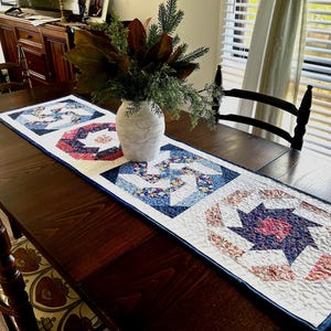 Op de afbeelding: Een witte tafelrunner met een blauw en rood patchwork ontwerp. De runner is gemaakt van vierkante stoffen met een geometrisch patroon. De runner ligt op een donkerhouten tafel met een vaas met groen in het midden.