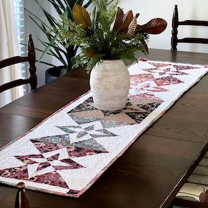 Peut inclure: Un chemin de table blanc avec un motif géométrique d'étoiles roses, brunes et bleues. Le chemin de table est sur une table en bois foncé avec un vase de verdure au centre.