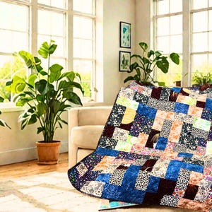 May include: A colorful patchwork quilt draped over a beige sofa. The quilt features a variety of patterned fabrics in shades of blue, brown, pink, and white. The room has large windows, potted plants, and a wooden chair.