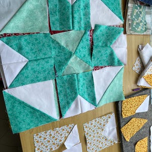 May include: A patchwork quilt block in progress, featuring a star design with white, teal, and yellow floral fabric pieces. The block is laid out on a wooden surface with additional fabric pieces and sewing tools nearby.