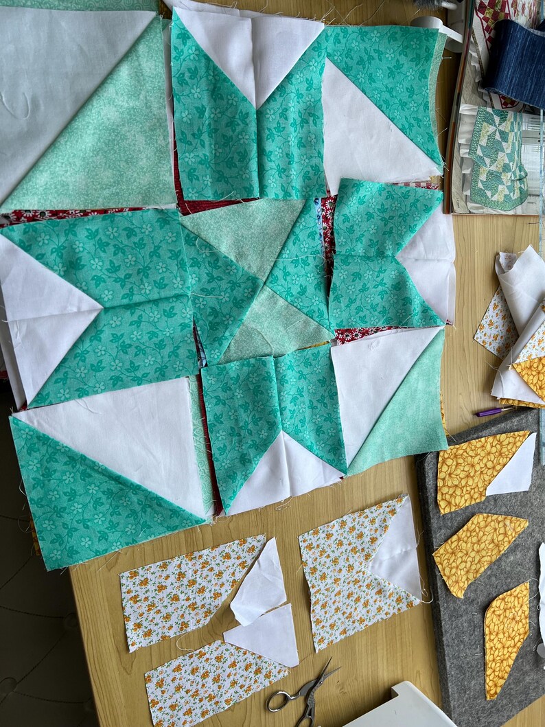 May include: A patchwork quilt block in progress, featuring a star design with white, teal, and yellow floral fabric pieces. The block is laid out on a wooden surface with additional fabric pieces and sewing tools nearby.