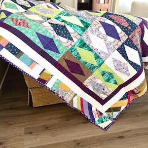 May include: A colourful quilt with a geometric pattern in shades of green, purple, brown, and white. The quilt is made of various fabrics with different patterns and textures. It is folded and draped over a brown sofa.