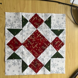 May include: A red and white patchwork quilt block with a star design. The fabric has a white background with snowflakes and gold lettering that says "peace", "hope", "joy", "light", and "hol".