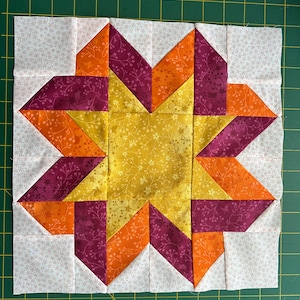 Puede incluir: Un bloque de colcha con un diseño de estrella en tonos naranja, amarillo y burdeos. La estrella está rodeada de un fondo blanco con un pequeño patrón floral.