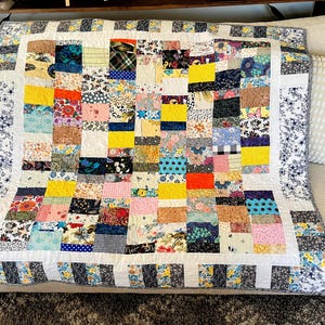 Op de afbeelding: Een patchwork quilt met een raster van veelkleurige stoffen vierkantjes in diverse patronen, waaronder bloemen, ruiten, stippen en effen kleuren. Het middengedeelte is omlijst door een witte rand en buitenpanelen van zwart-witte bloemenstof.