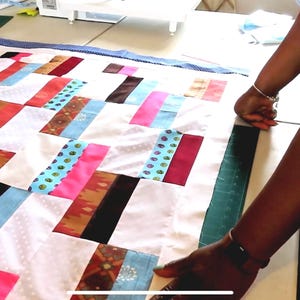 May include: A person is working on a quilt made of colorful fabric squares. The quilt is laid out on a table and the person is smoothing out the fabric. The quilt is made of a variety of colors and patterns, including pink, blue, green, and brown.