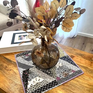 Puede incluir: Un arreglo decorativo de flores y hojas secas en un jarrón de cristal transparente, colocado sobre un mantel individual con estampado de patchwork. El jarrón está sobre una mesa de centro de madera, con un álbum de fotos blanco al fondo.