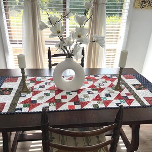 Può includere: Un runner da tavolo in patchwork rosso, bianco e blu con sfondo bianco e un motivo geometrico. Il runner è su un tavolo in legno scuro con due candelieri bianchi e un vaso bianco con fiori bianchi.