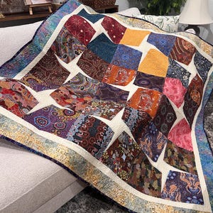 May include: A patchwork quilt featuring a variety of square fabric blocks in shades of red, orange, yellow, blue, and purple, separated by cream-colored sashing. The quilt has a decorative border with a blue and gold mottled pattern.