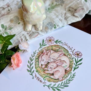 May include: A watercolor illustration of three brown bunnies sleeping in a nest surrounded by green leaves and pink flowers.