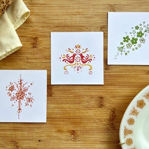 May include: Three small cards with different folk art designs. The first card has a red and yellow design with two birds facing each other. The second card has a green design with a cluster of flowers. The third card has a red and yellow design with a flower and butterfly.