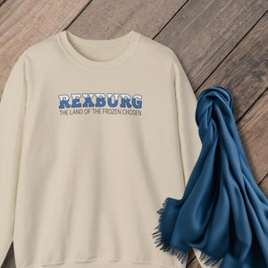 May include: A cream-colored sweatshirt with a blue and white graphic that says "Rexburg, The Land of the Frozen Chosen".
