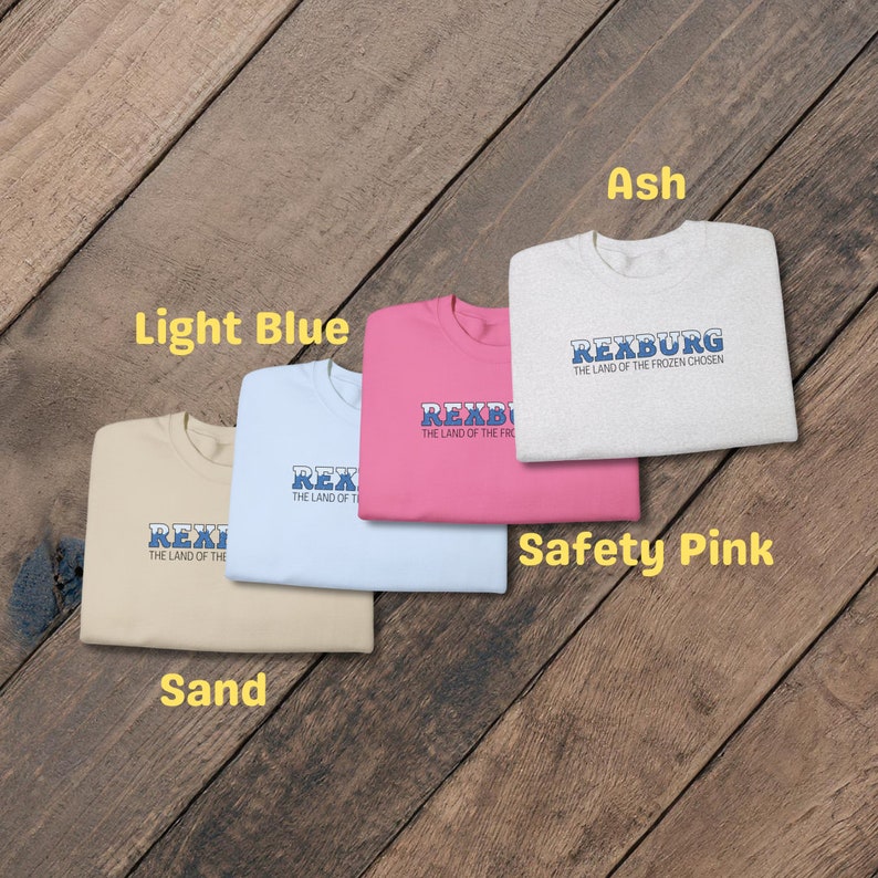 May include: Five t-shirts in different colors, each with the same design. The design features the text "REXBURG" in blue and white letters, with the words "THE LAND OF THE FROZEN CHOSEN" underneath. The t-shirts are in the colors ash, light blue, safety pink, sand, and white.
