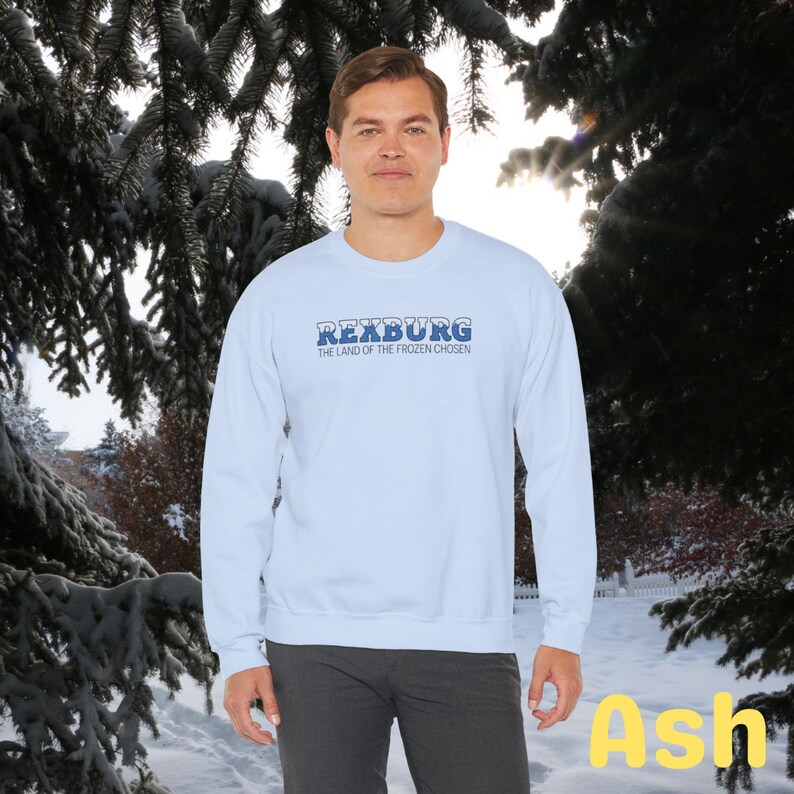 May include: A light blue sweatshirt with the text "Rexburg, The Land of the Frozen Chosen" in blue lettering. The sweatshirt is being modeled by a person against a snowy background.