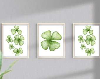 May include: Three framed watercolor prints featuring green shamrocks. The prints are displayed on a gray wall. Each print has a light wooden frame. The shamrocks vary in size and arrangement, with one print showcasing a four-leaf clover.