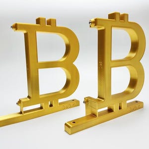 May include: Two gold-colored, 3D-printed Bitcoin symbol bookends standing upright. Each piece features the letter B with two vertical lines and the text 3w3.io embossed on the base. The objects have a smooth, metallic finish and a flat, rectangular base with visible screw holes for mounting.
