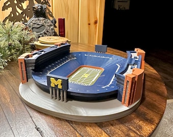 Può includere: Un modello dettagliato di uno stadio di football, con un campo blu con linee di demarcazione e la parola "MICHIGAN" in giallo. Lo stadio ha aree di seduta blu, accenti arancioni e grigi e un grande logo "M". Il modello è su una superficie di legno.