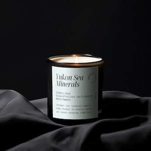 May include: A black candle jar with a white label that reads "Yukon Sea Minerals" and lists the scent notes: Ozone | Linen | Eucalyptus | Sea Salt | Freesia | Moss | Powder. The label also states that the candle is a coconut soy scented candle, hand poured in Myrtle Beach, and has a 48-hour burning time.