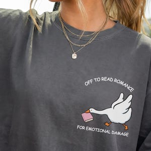 Puede incluir: Camiseta color pimienta con un dibujo de un ganso de dibujos animados llevando un libro. El texto blanco dice: "OFF TO READ ROMANCE FOR EMOTIONAL DAMAGE". La camiseta es de corte holgado, ideal para uso diario.