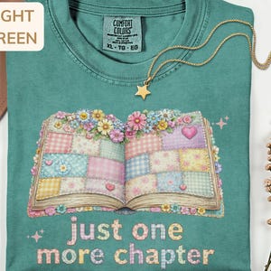 Puede incluir: Camiseta verde claro con un gráfico de un libro abierto decorado con flores y cuadrados de patchwork. El texto "just one more chapter" está debajo del libro. Un collar de estrella dorada está sobre la camiseta.