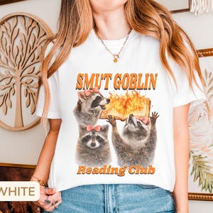 May include: White t-shirt with the text "SMUT GOBLIN READING CLUB" in orange. The graphic features three raccoons, each wearing a pink bow, with one holding a flaming book. The shirt is a casual, humorous design.