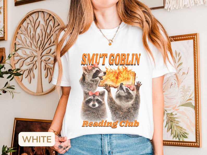 May include: White t-shirt with the text "SMUT GOBLIN READING CLUB" in orange. The graphic features three raccoons, each wearing a pink bow, with one holding a flaming book. The shirt is a casual, humorous design.