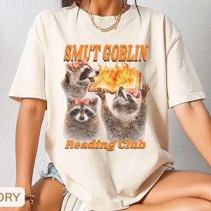 May include: Ivory-colored t-shirt featuring a graphic of three raccoons with pink bows, holding a book with flames. The text "SMUT GOBLIN" and "Reading Club" are in orange. The shirt is a casual, short-sleeved style.