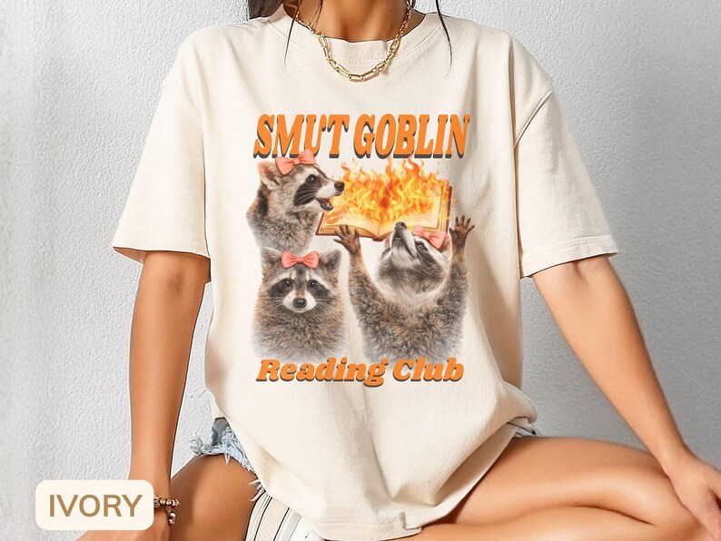 May include: Ivory-colored t-shirt featuring a graphic of three raccoons with pink bows, holding a book with flames. The text "SMUT GOBLIN" and "Reading Club" are in orange. The shirt is a casual, short-sleeved style.