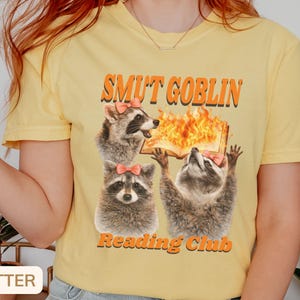 May include: Butter-colored t-shirt featuring a graphic of three raccoons with pink bows, a burning book, and the text "SMUT GOBLIN Reading Club" in orange. The shirt is a casual, crew neck style.