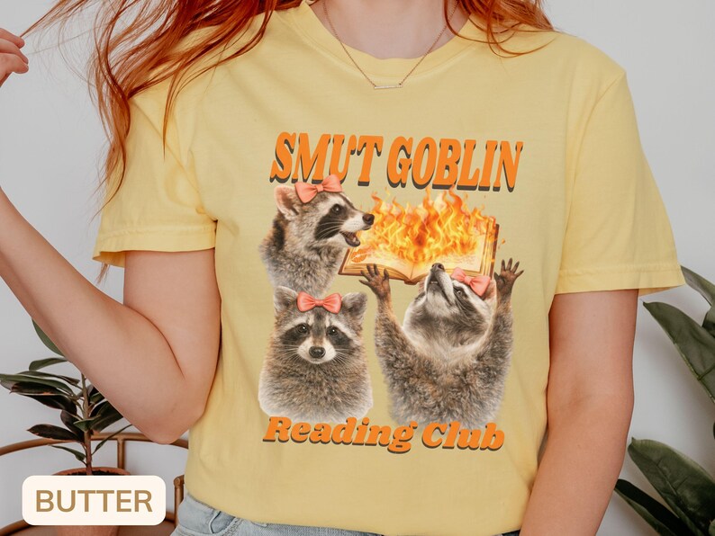 May include: Butter-colored t-shirt featuring a graphic of three raccoons with pink bows, a burning book, and the text "SMUT GOBLIN Reading Club" in orange. The shirt is a casual, crew neck style.