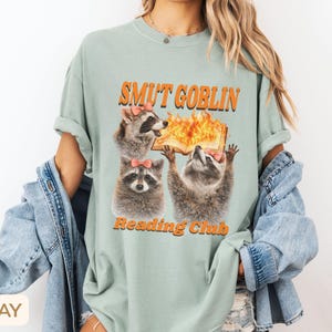 May include: Sage green t-shirt with the text "SMUT GOBLIN Reading Club" in orange. The graphic features three raccoons with pink bows, one holding a flaming book. The shirt has short sleeves and a relaxed fit.