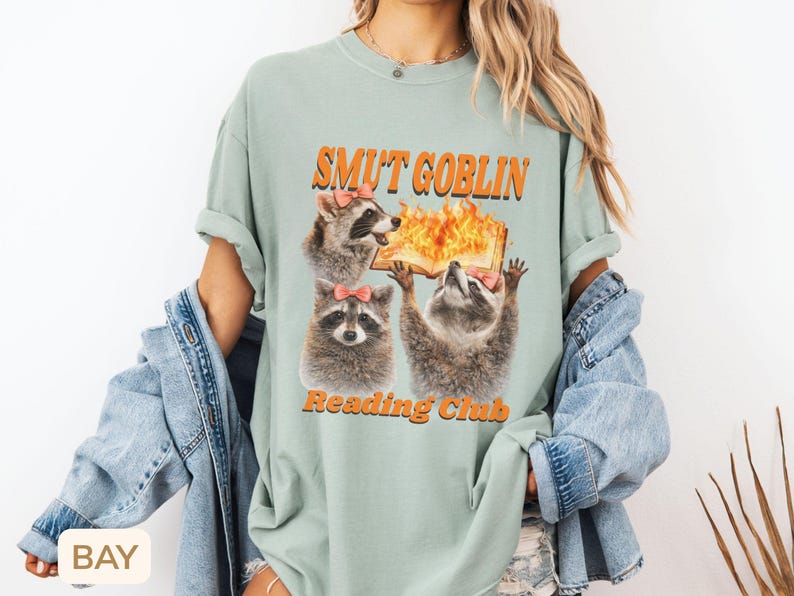 May include: Sage green t-shirt with the text "SMUT GOBLIN Reading Club" in orange. The graphic features three raccoons with pink bows, one holding a flaming book. The shirt has short sleeves and a relaxed fit.