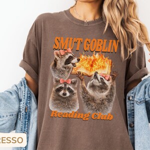 May include: Espresso-colored t-shirt featuring a graphic of three raccoons wearing pink bows. The text "SMUT GOBLIN Reading Club" is printed above and below the raccoons, with a burning book graphic.