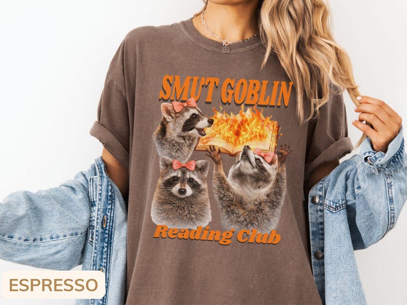 May include: Espresso-colored t-shirt featuring a graphic of three raccoons wearing pink bows. The text "SMUT GOBLIN Reading Club" is printed above and below the raccoons, with a burning book graphic.