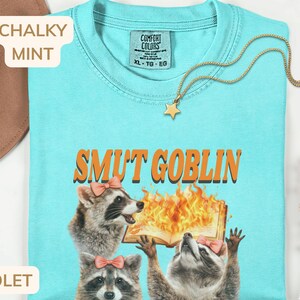 May include: A light blue t-shirt with the text "SMUT GOBLIN" above a graphic of raccoons and a burning book. The shirt has a crew neck and short sleeves. A gold star pendant necklace is draped across the shirt.