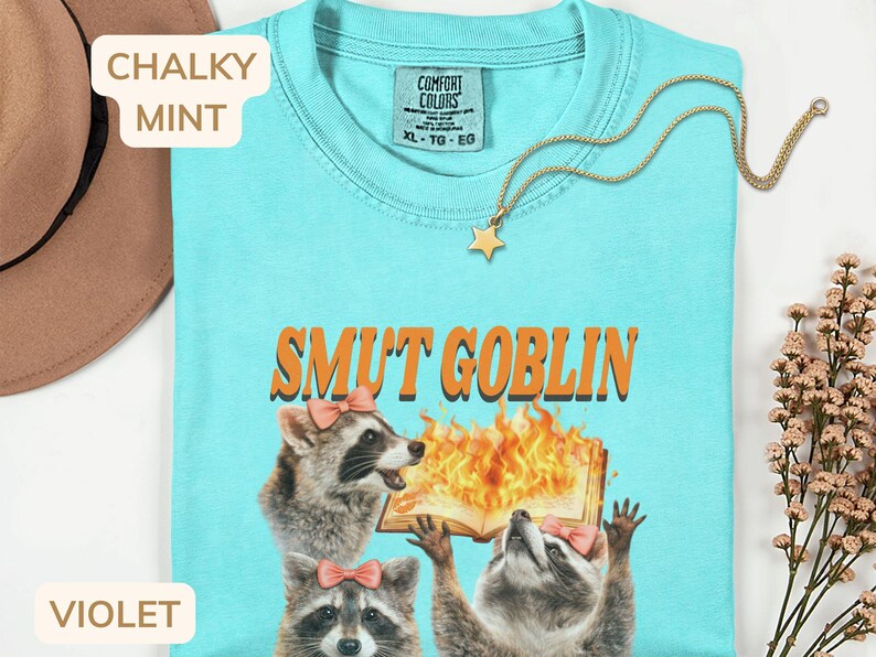 May include: A light blue t-shirt with the text "SMUT GOBLIN" above a graphic of raccoons and a burning book. The shirt has a crew neck and short sleeves. A gold star pendant necklace is draped across the shirt.