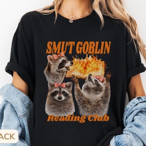 May include: Black t-shirt with the text "SMUT GOBLIN Reading Club" in orange. The graphic features three raccoons with pink bows, one holding a flaming book. The shirt is a casual, unisex style.