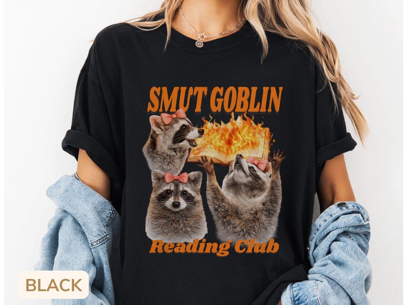 May include: Black t-shirt with the text "SMUT GOBLIN Reading Club" in orange. The graphic features three raccoons with pink bows, one holding a flaming book. The shirt is a casual, unisex style.